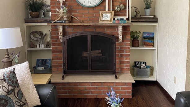 A living room with a brick fireplace, a large clock, and shelves with decorative items.