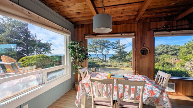 Una habitación de madera con una mesa y sillas, ventanas amplias y vistas a árboles y al mar.