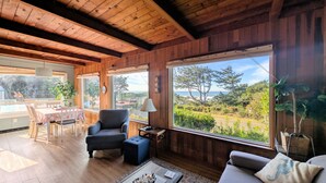 Una habitación acogedora con paredes y techo de madera, una zona de comedor con mesa y sillas, un sillón cómodo, una mesita auxiliar con lámpara, y amplios ventanales que ofrecen vistas al exterior.