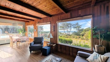 Une pièce chaleureuse aux murs et au plafond en bois, un coin repas avec une table et des chaises, un fauteuil confortable, une petite table d’appoint avec une lampe, et de grandes fenêtres offrant une vue sur l’extérieur.