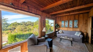 Una sala de estar acogedora con techo de madera, ventanas amplias que dan a vistas de vegetación, un sofá gris, una mesita y una lámpara.