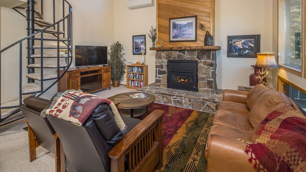 A cozy living room with a fireplace, a stone fireplace surround, a comfortable leather sofa, a wooden coffee table, a bookshelf, and a spiral staircase. - Beautiful home w/fireplace, amazing views & community hot tub - Hidden Village Cozy Getaway (Gallatin Gateway)