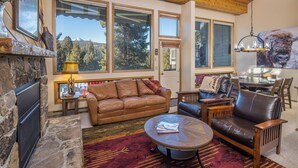 A cozy living room with a stone fireplace, leather furniture, large windows with mountain views, and a dining area with a chandelier. - Beautiful home w/fireplace, amazing views & community hot tub - Hidden Village Cozy Getaway (Gallatin Gateway)