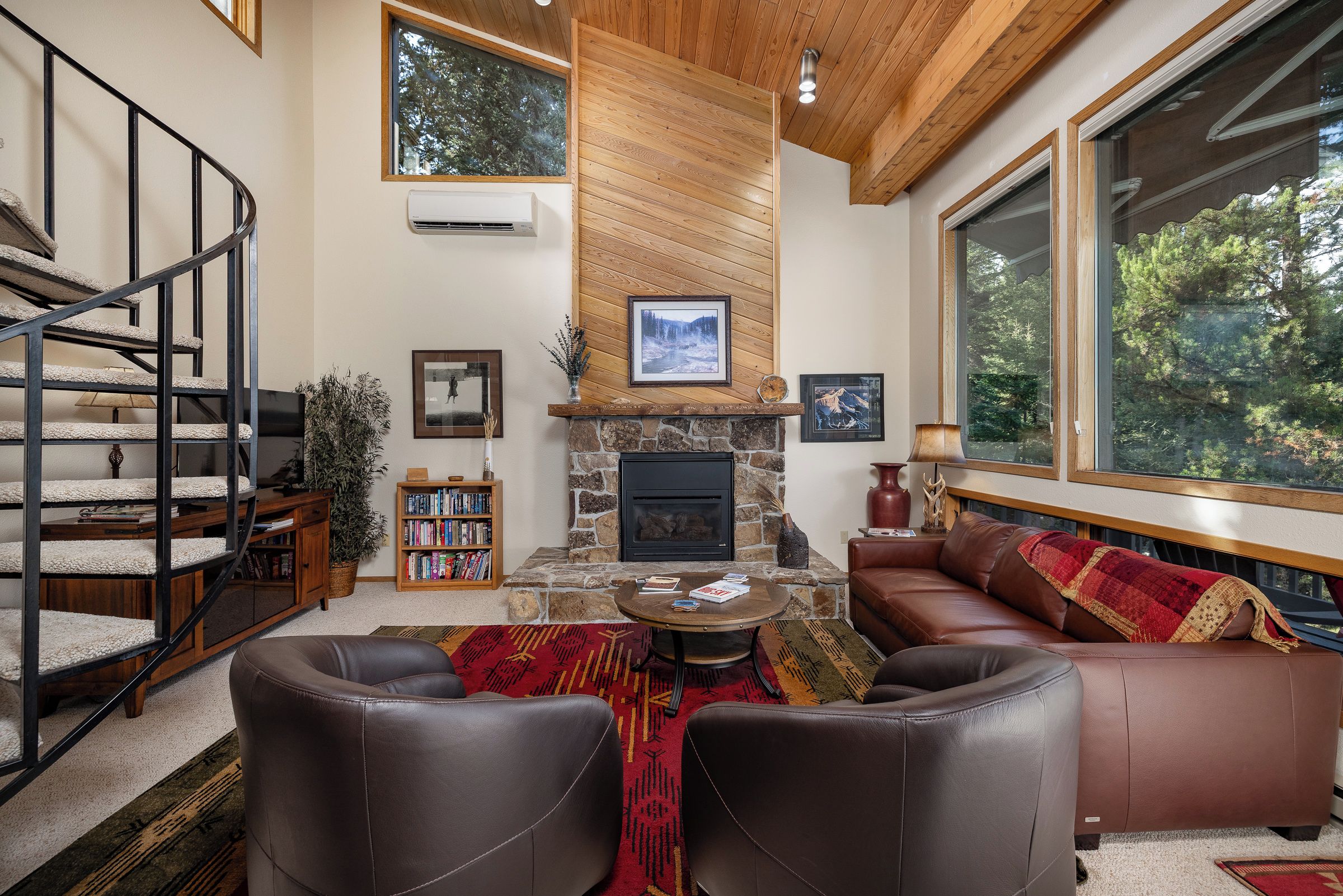 A cozy living room with a stone fireplace, a staircase, and a large window overlooking trees.
