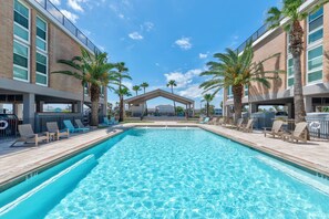 Una piscina con sillones de descanso y palmeras, rodeada de edificios con ventanales amplios y una zona cubierta al fondo.