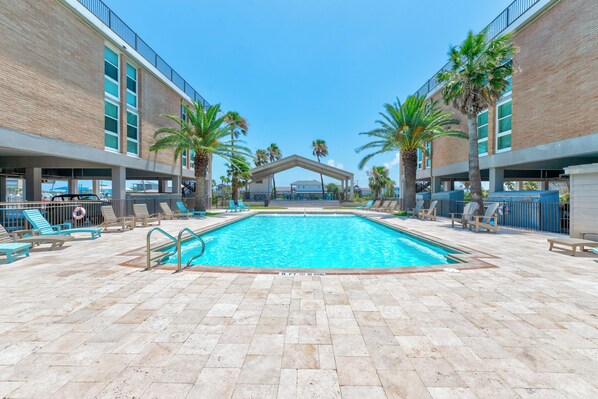 Una piscina con sillones de descanso y palmeras, rodeada de edificios con fachadas de ladrillo y ventanas grandes.
