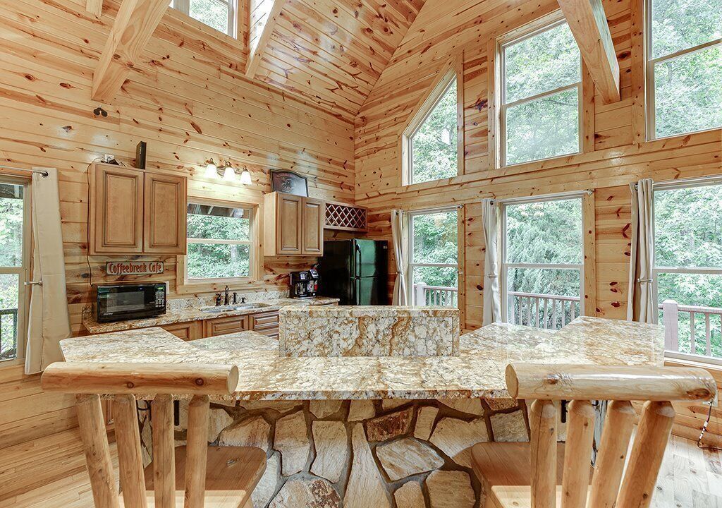 Una cabaña con base de piedra, paredes de madera, una cocina con encimera de piedra y amplias ventanas que ofrecen vistas del bosque circundante.