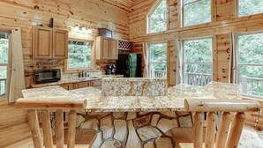 Une cabane avec des fondations en pierre, des murs en bois, une cuisine dotée d’un plan de travail en pierre et de grandes fenêtres offrant une vue sur la forêt environnante.