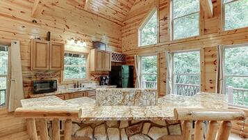 Una cabaña con base de piedra, paredes de madera, una cocina con encimera de piedra y amplias ventanas que ofrecen vistas del bosque circundante.