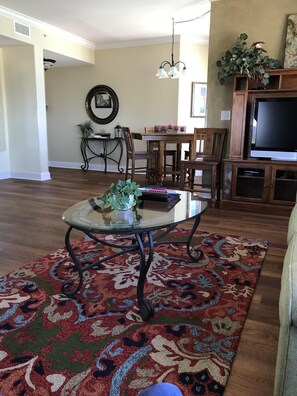 Una sala de estar con una mesa de centro de vidrio, un tapete estampado, piso de madera, una zona de comedor con mesa y sillas, y un televisor sobre un mueble de madera.