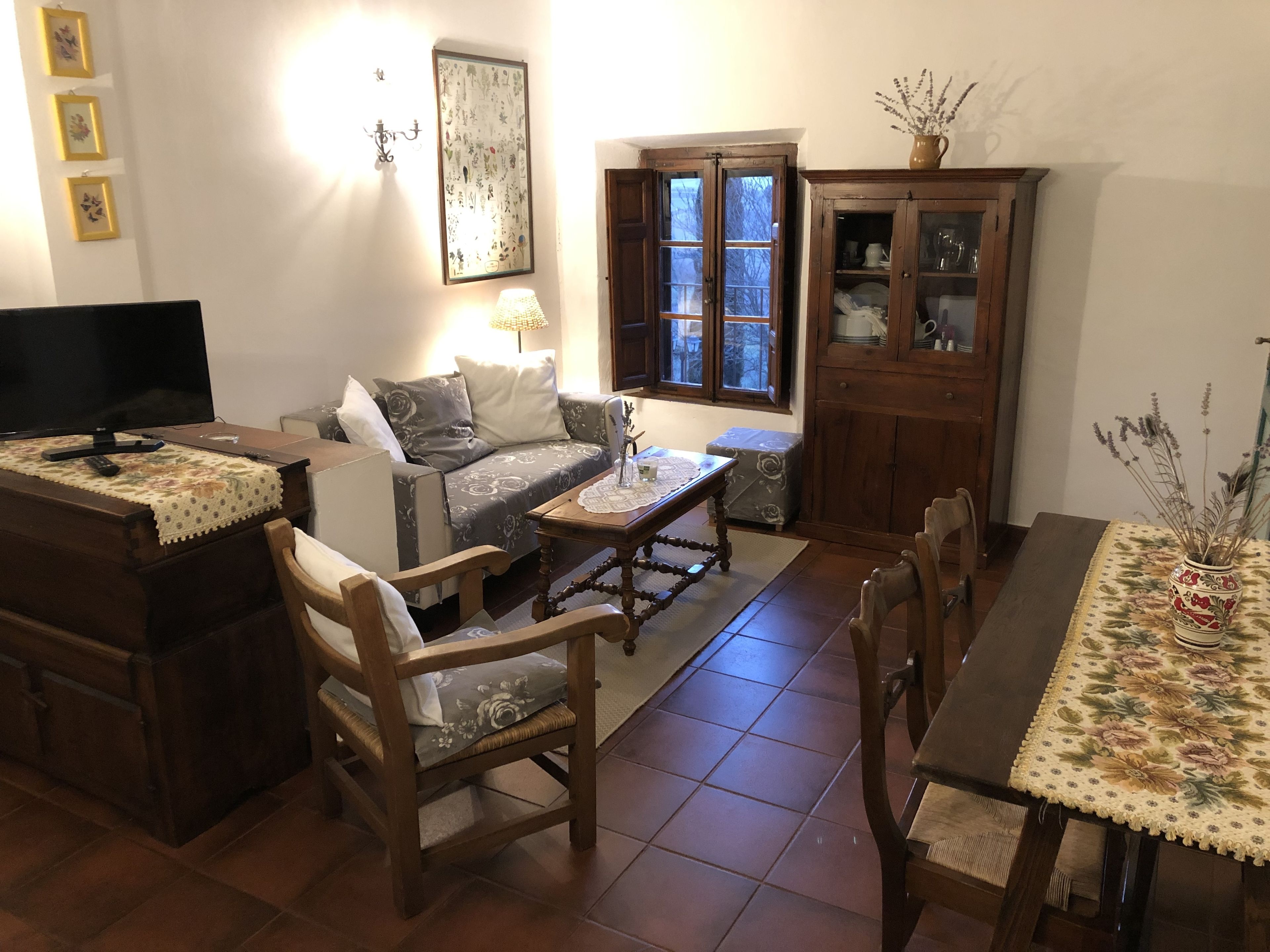 Een woonkamer met een bank, twee houten stoelen, een salontafel, een televisie, een kast en een raam met uitzicht op bomen.