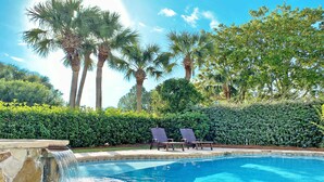 A swimming pool with a stone waterfall, surrounded by palm trees and a hedge.