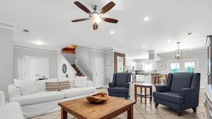 A spacious living room with a wooden coffee table, white sofa, and blue armchairs. A ceiling fan and kitchen are visible in the background.