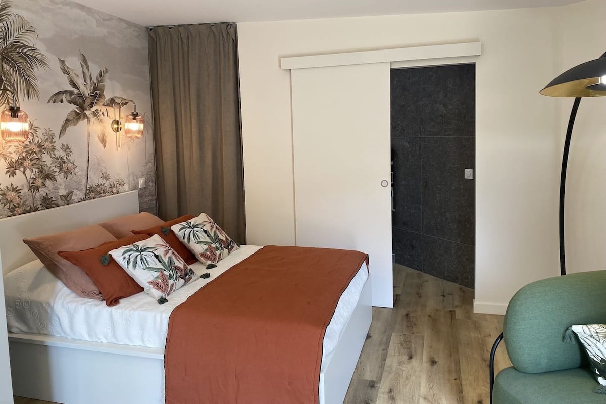 Une chambre à coucher avec un lit, une table de chevet, une lampe fixée au mur, une porte et un fauteuil vert.
