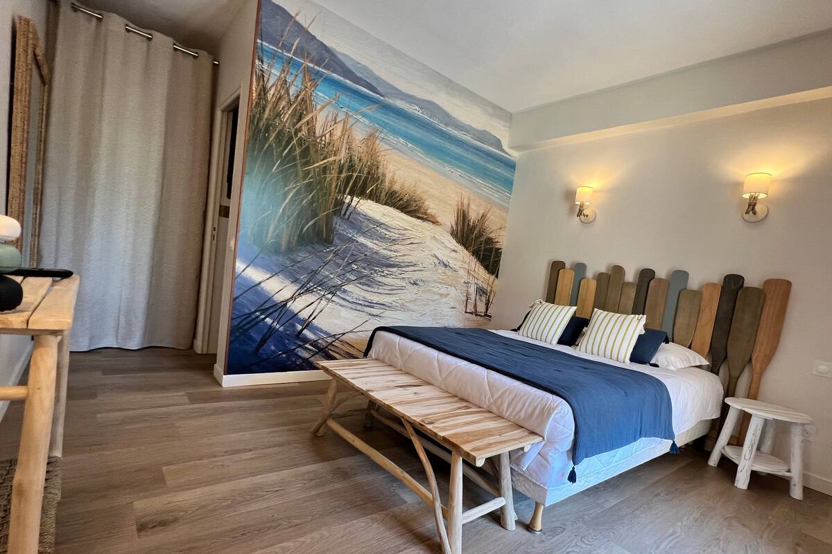 Une chambre à coucher comprenant un lit, des tables de chevet, un banc, un tableau représentant une scène de plage et deux lampes fixées au mur.