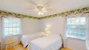A bedroom with two single beds, a ceiling fan, a nightstand with a lamp, a chair, and windows with blinds. - Grace Cottage 3 bedrooms, 2 bathrooms (Jekyll Island)