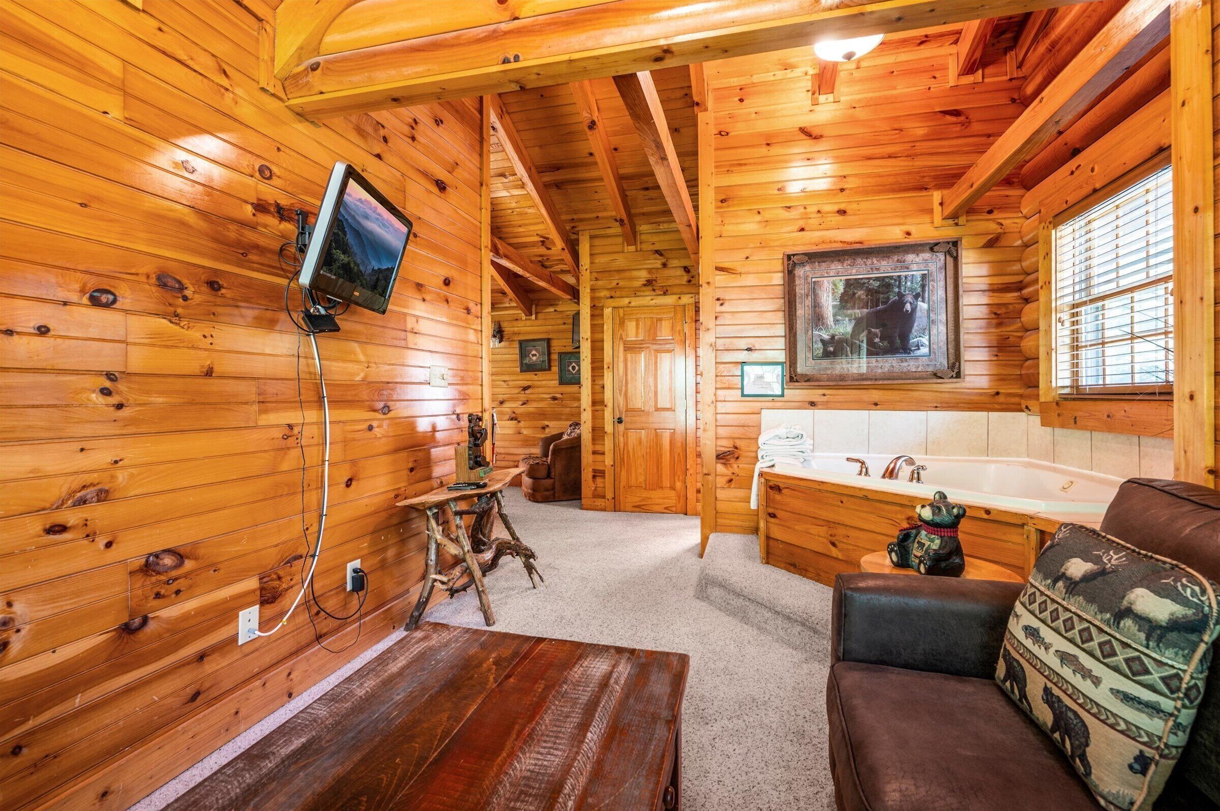 Interior de una cabaña de troncos con detalles de madera, un televisor montado, una bañera y una zona de estar acogedora.