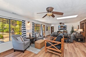 A spacious living room with a large window, a ceiling fan, and a television.