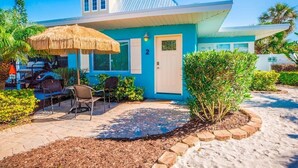 Una casa moderna con facciata blu, il numero '2' sulla porta, un patio con sedie e un ombrellone di paglia.