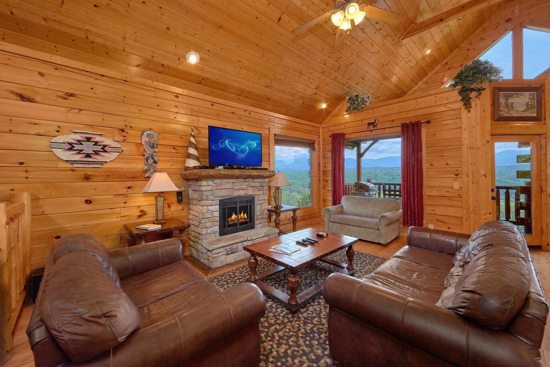A cozy log cabin interior with a stone fireplace, leather sofas, a flat-screen TV, and a scenic mountain view.