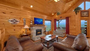 Acogedor interior de cabaña de madera con chimenea de piedra, sofás de cuero, televisor de pantalla plana y vistas panorámicas a la montaña.
