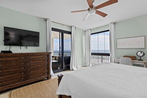 A bedroom with a bed, a dresser, a television, a ceiling fan, and a view of the beach.