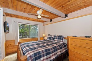 Un dormitorio con techo de madera, una cama con ropa de cama escocesa, un aparador de madera, una mesita de noche, una lámpara y un televisor en la pared.