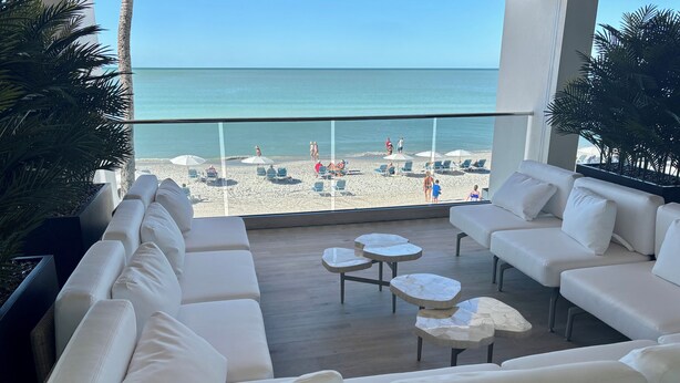 Un balcon avec des canapés blancs et des tables rondes donnant sur une plage parsemée de parasols et où se trouvent des gens.