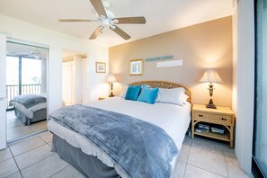 A bedroom with a large bed, two bedside tables with lamps, a ceiling fan, and a view of the beach.