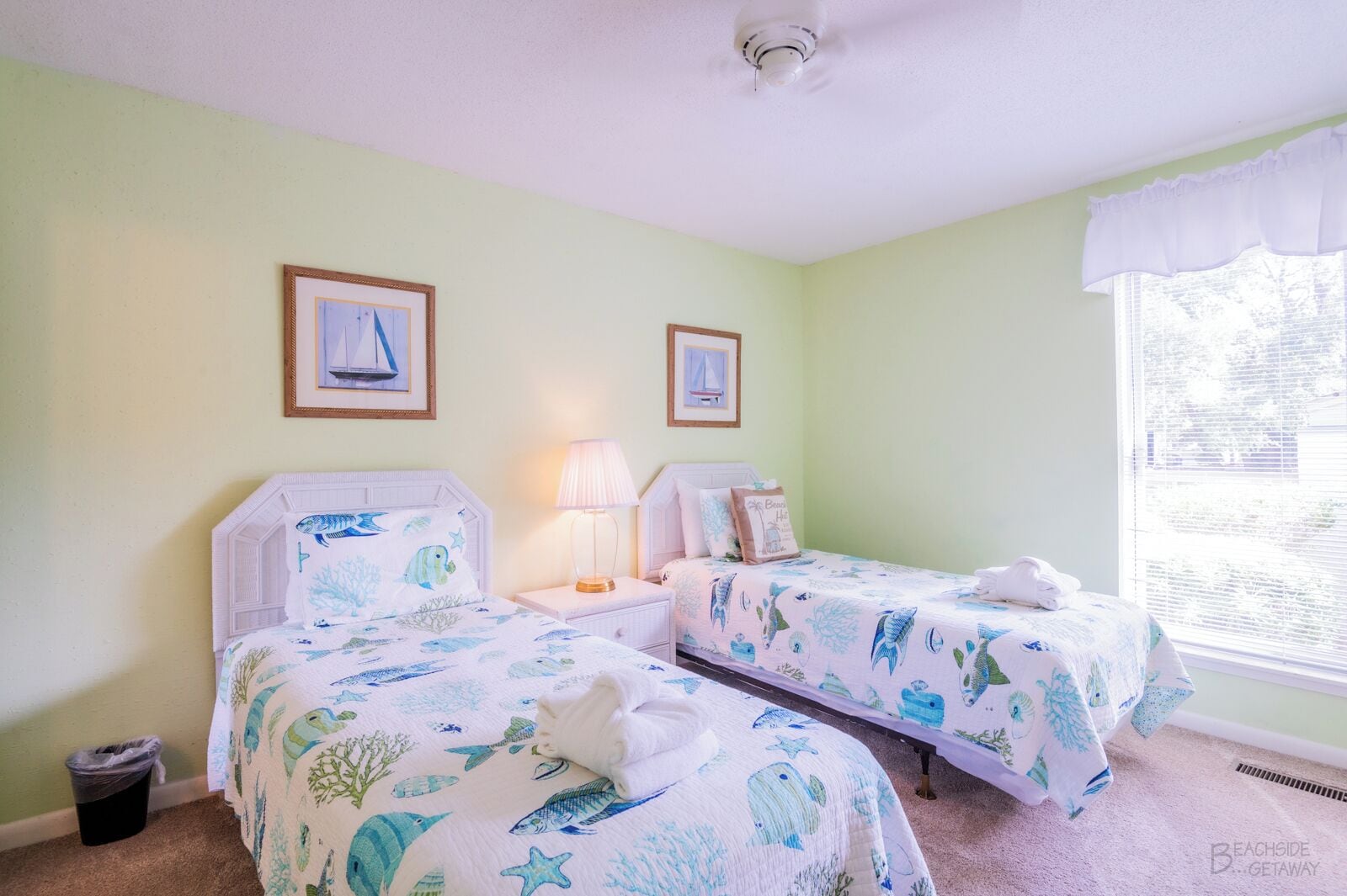 A room with two beds, each with a nautical-themed bedspread, a bedside lamp, and framed pictures on the wall.