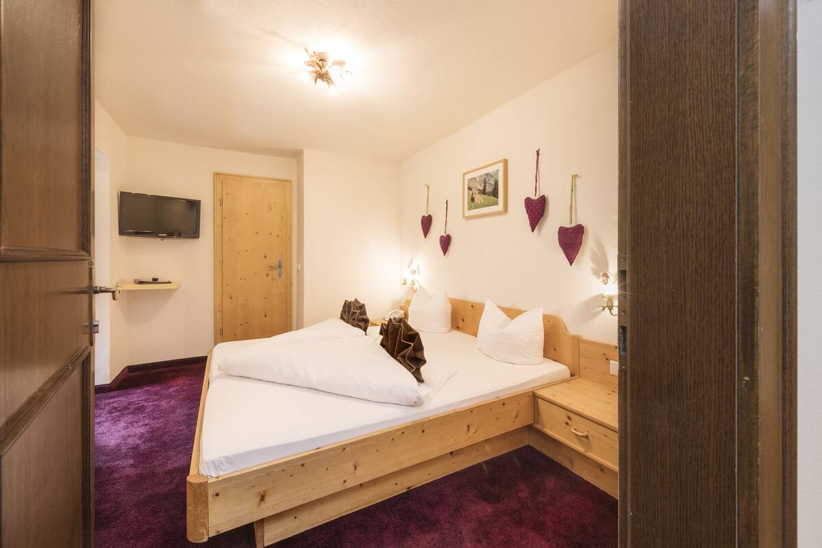 A wooden bed with white linens, a wooden nightstand, a television on the wall, and heart-shaped decorations.