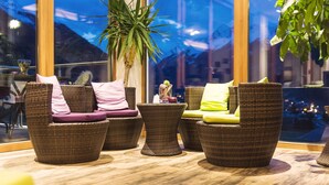 A seating area with wicker chairs and cushions, a potted plant, and a view through large windows. - Lifestyle Panorama Hotel Erika (Neustift Im Stubaital)