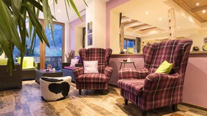 A cozy living room with plaid armchairs, a round ottoman, and a view of a deck. - Lifestyle Panorama Hotel Erika (Neustift Im Stubaital)
