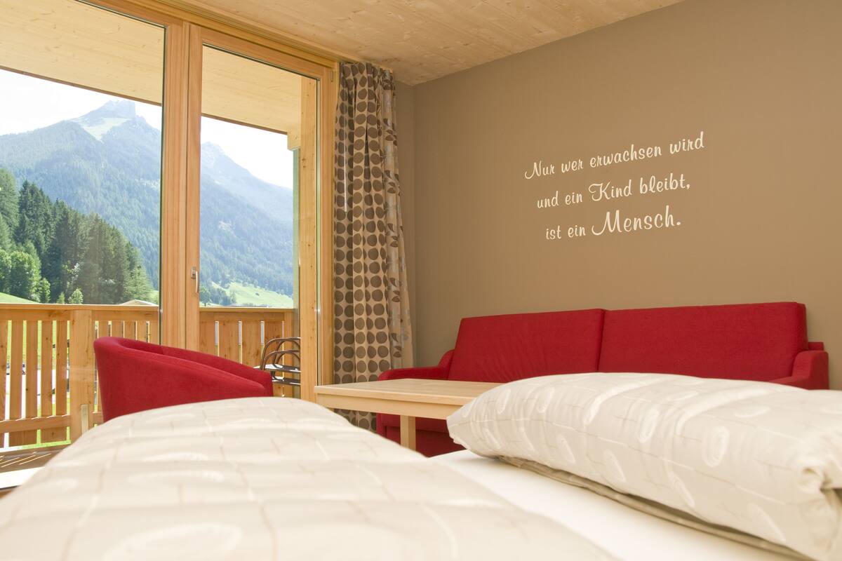 A bedroom with a wooden ceiling, a red sofa, a wooden table, and a large window overlooking mountains.