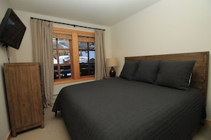 Ein Schlafzimmer mit Bett, Holz-Kopfteil, Nachttisch, Lampe, einem an der Wand montierten Fernseher und einem Fenster mit Vorhängen.