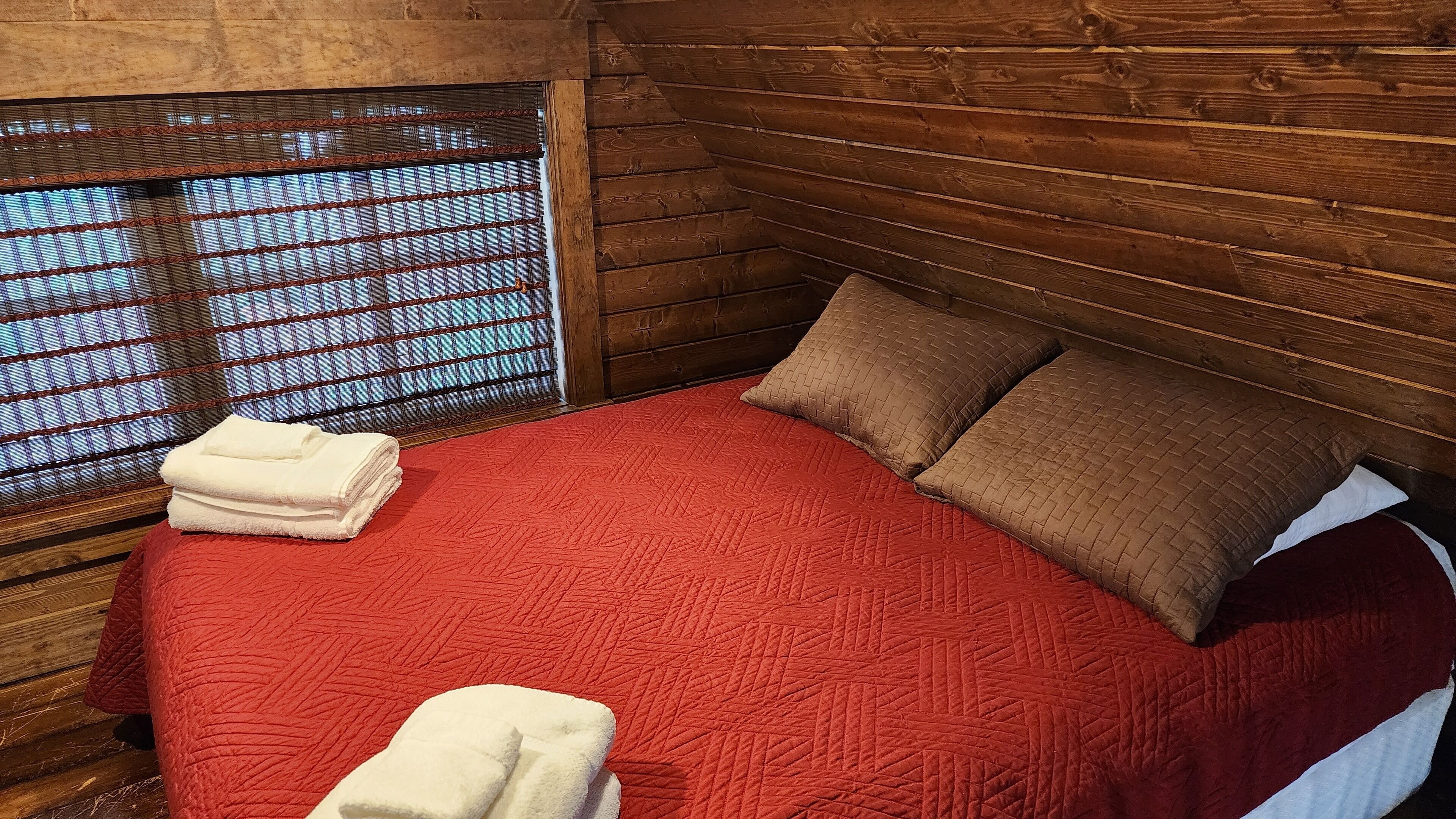 A wooden cabin room with a red quilted bedspread, two pillows, and a window with a grid pattern.