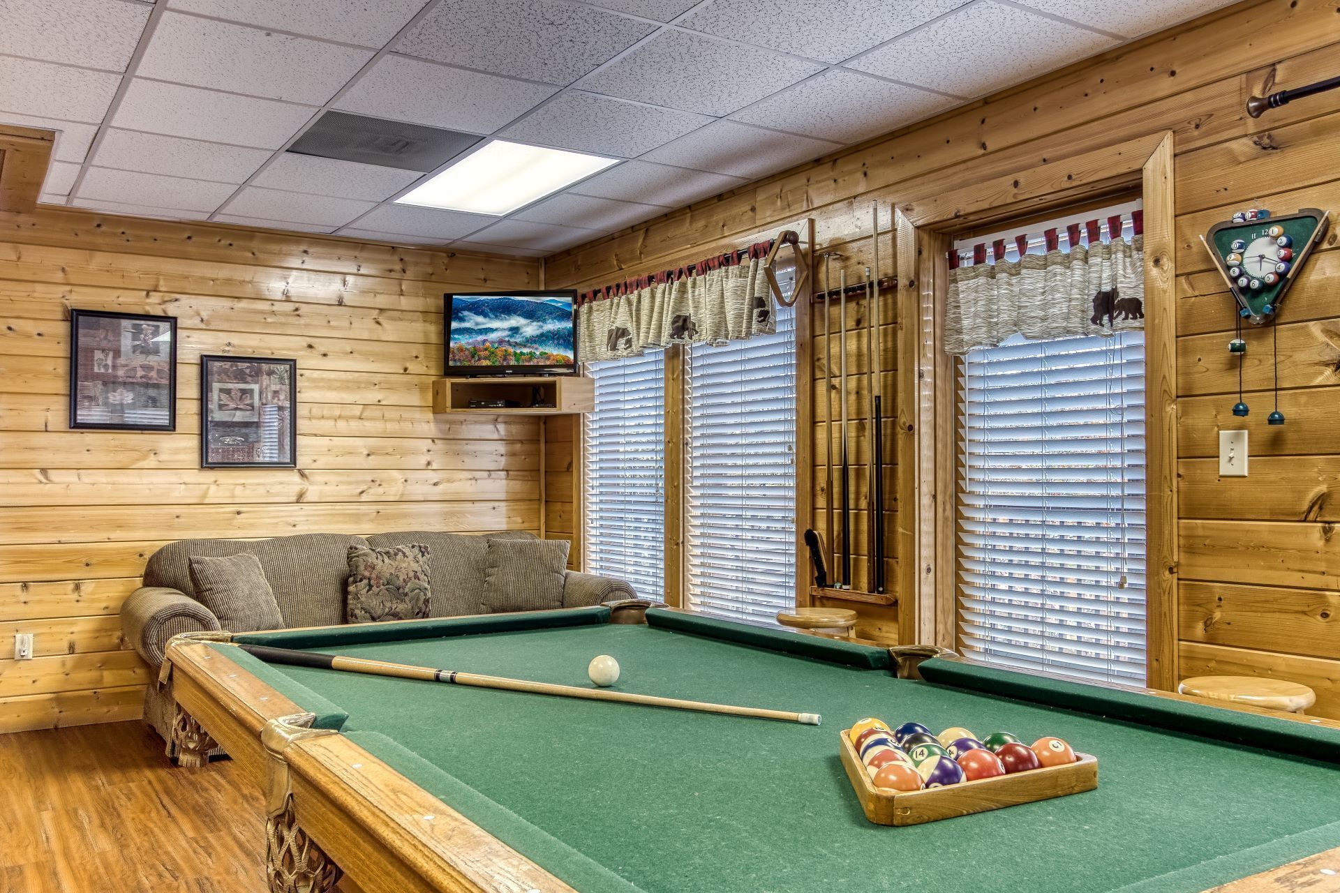 A pool table with a cue stick and balls, a sofa, wooden walls, a framed picture, and a clock.