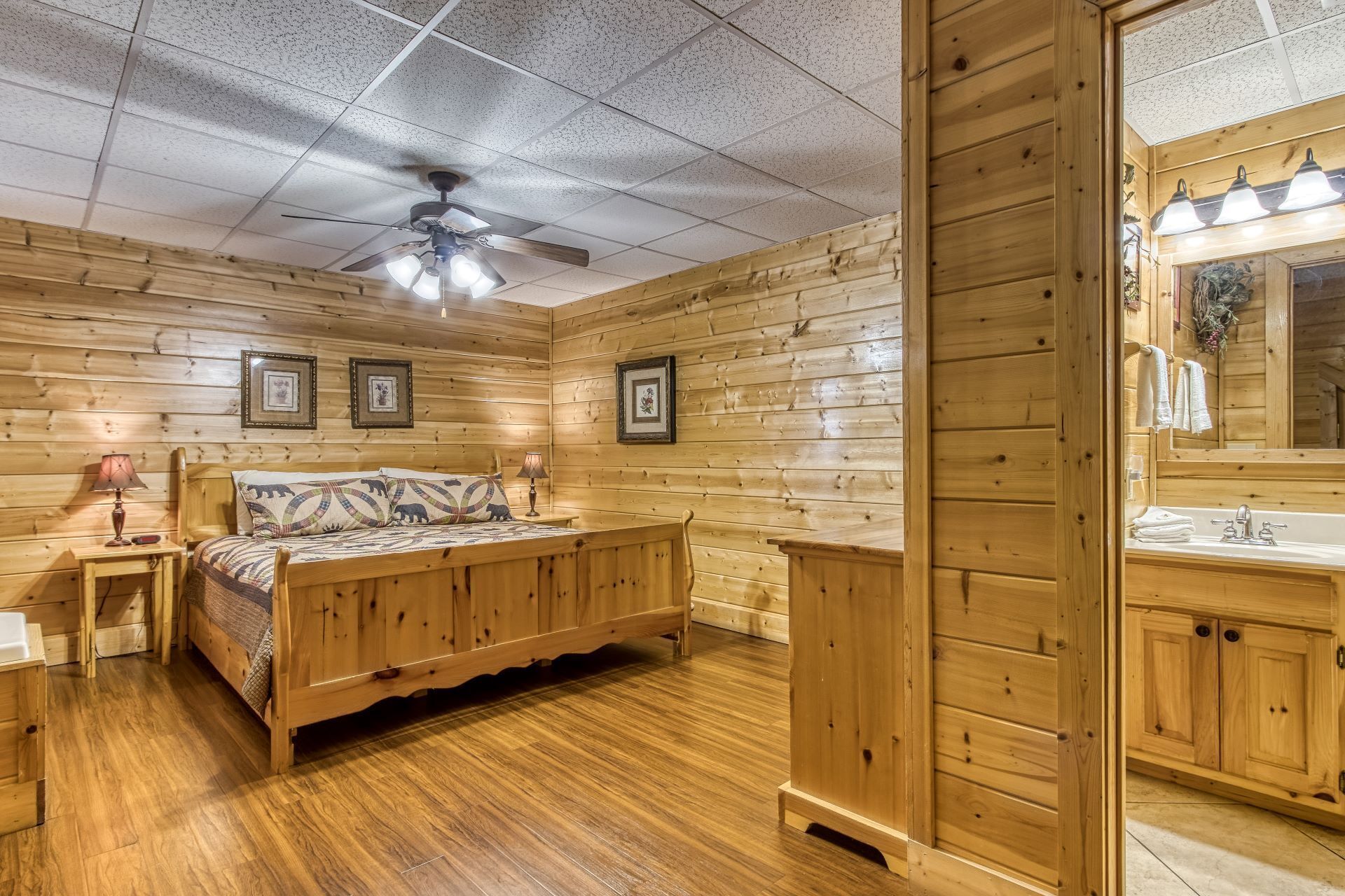 A wooden cabin room with a bed, bedside tables, a ceiling fan, framed pictures, and a bathroom with a sink and mirror.