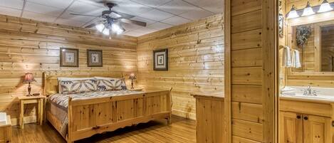 A wooden cabin room with a bed, bedside tables, a ceiling fan, framed pictures, and a bathroom with a sink and mirror.