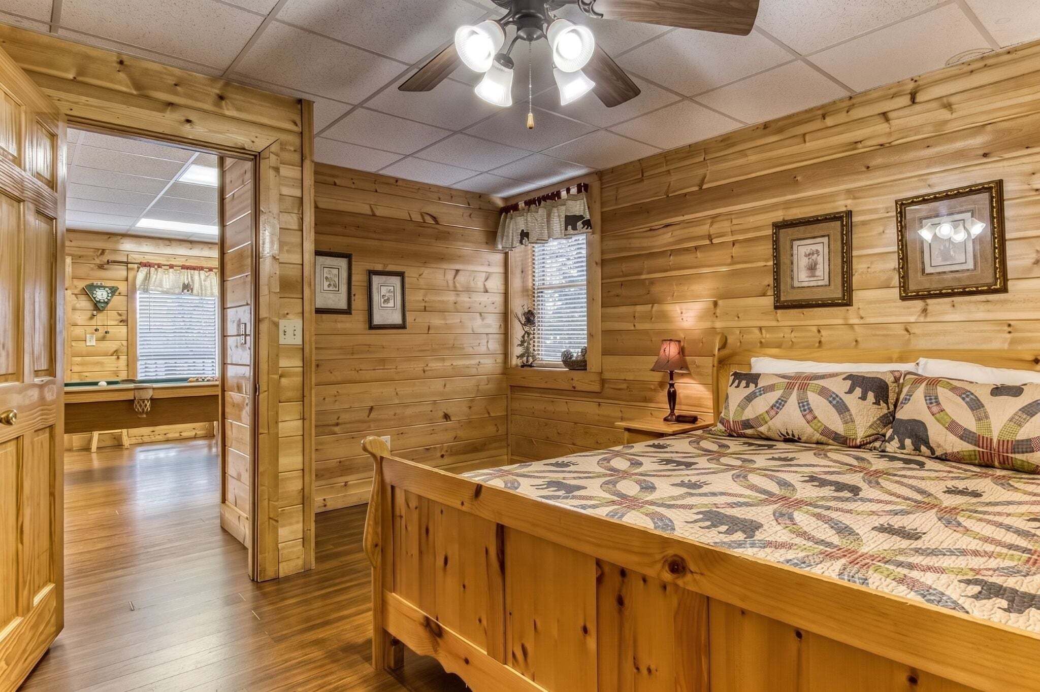 A wooden cabin interior with a bed, a ceiling fan, framed pictures, and a window with blinds.