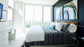 A modern bedroom with a large bed, a desk, and a chair. There is a large window providing a view of the city. - Coconut Grove 21st Floor Private Studio-Parking & WIFI included (Miami)