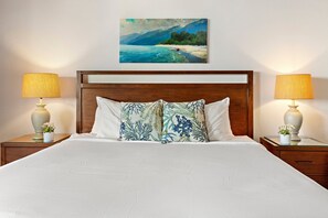Un lit bien fait, avec une tête de lit en bois, deux tables de chevet avec des lampes et un tableau représentant un paysage côtier au-dessus du lit.