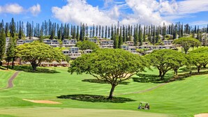 Un campo de golf con un club de campo, rodeado por un bosque de árboles altos y una hilera de edificios residenciales al fondo.