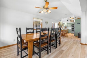 Een eetruimte met een houten tafel en stoelen, een plafondventilator, en een keuken op de achtergrond.