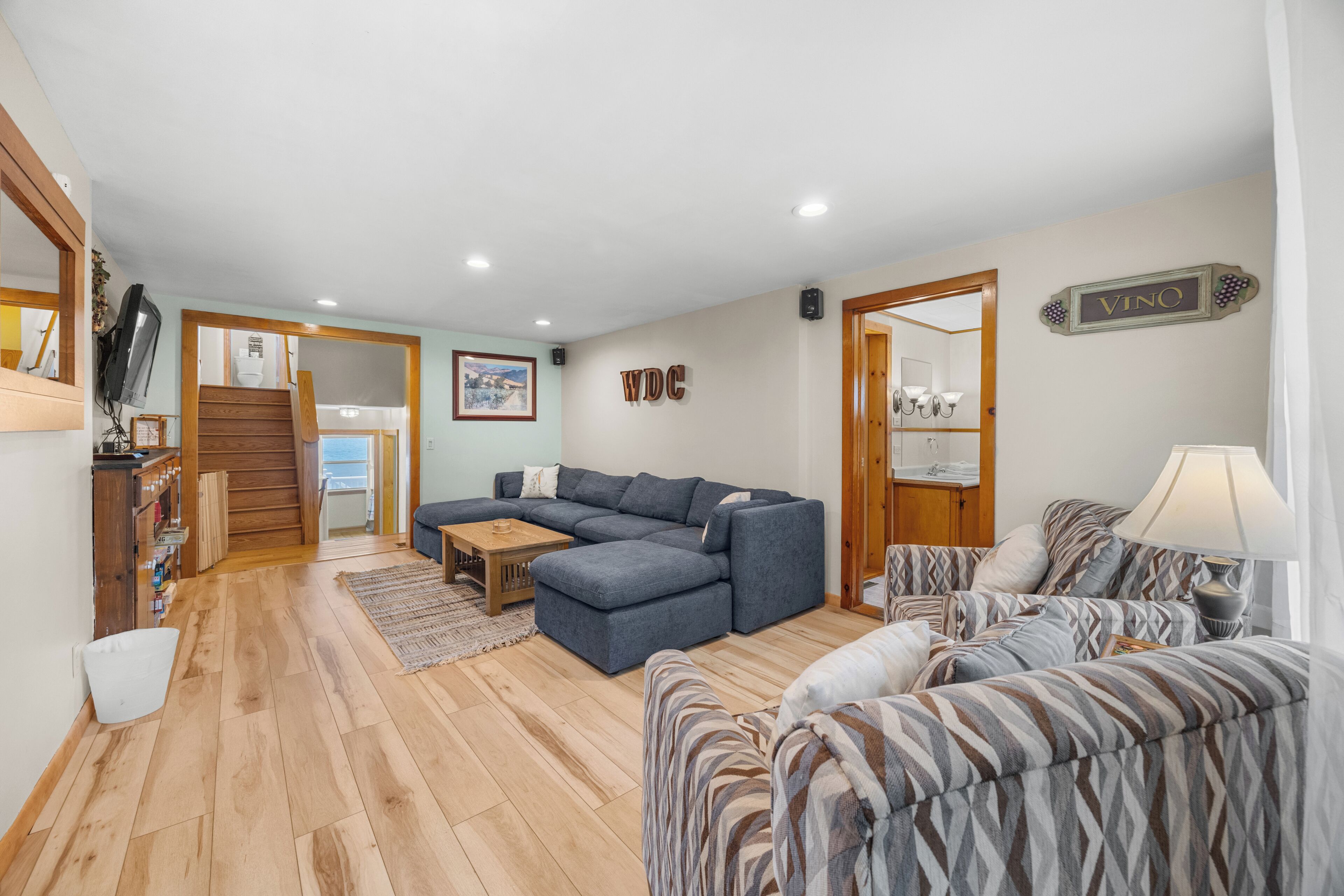 A spacious living area with a large sectional sofa, a patterned armchair, and a wooden coffee table. A staircase leads to an upper level, and a painting is visible on the wall.