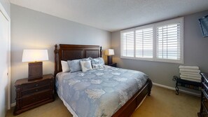A bedroom with a large bed, two bedside tables with lamps, a window with blinds, and a chair with folded towels. - The Islander 704 - Complimentary Beach (Seasonally) - Heated Pool! (Destin)