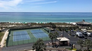 Tenniskenttäalue, jolta on näkymä rannalle, ja jolla on myös pysäköintialue ja pieni katos.