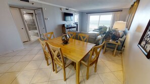 A dining area with a wooden table and chairs, a television above the table, and a view of the ocean through large windows. - The Islander 704 - Complimentary Beach (Seasonally) - Heated Pool! (Destin)