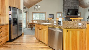 A kitchen with wooden cabinets, a stainless steel refrigerator, a dishwasher, granite countertops, and a stone fireplace. - TIMB03 - Classic Lodge Style Home, Single Level, Ping Pong & Foosball, Hot Tub (Sunriver)
