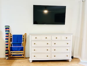 Un tocador blanco con pomos de latón, una silla azul con cojín a rayas y un televisor de pantalla plana montado en la pared.